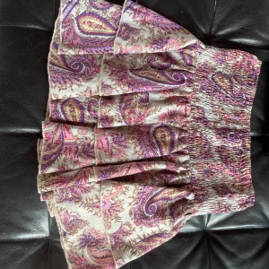 Mönstrad kortkjol i rosa och lila - Supersöt kortkjol med mönster i rosa, lila och beige toner. Kjolen har bred smock i midjan för en bekväm passform och volangig nederdel. Köpt i Spanien för 600🥰