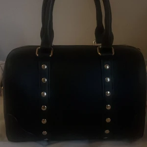 Svart handväska med nitar - Cool svart handväska i skinnimitation med två rejäla handtag och snygga guldfärgade nitar framtill. Väskan har en stilren, rundad form och passar perfekt för dig som gillar en edgy look.