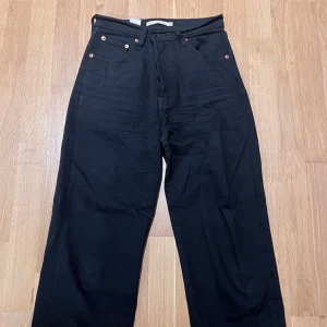 Levis - Svarta raka jeans med bred passform - Säljer ett par svarta jeans med raka ben och bred passform. Klassisk femficksmodell med knappgylf och detaljer i kopparfärg. Strl: 26x27