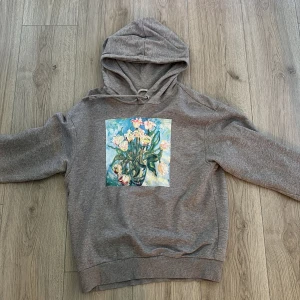 Grå hoodie med blommigt tryck från Monki - Grå hoodie från Monki med ett stort färgglatt blommotiv på bröstet. Tröjan har huva med snörning och lång ärm. 