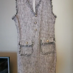 Rutig ärmlös kortklänning med pärlknappar - Snygg ärmlös kortklänning i rutigt tweedtyg med fransade kanter och dekorativa pärlknappar framtill. Klänningen har två fickor med lock och pärldetaljer. Färgerna går i vitt, svart och lite rosa. Perfekt för dig som gillar en chic och trendig look.