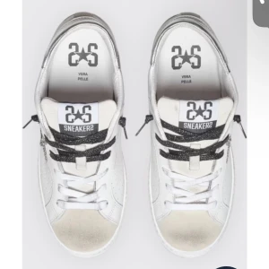 Vita sneakers från 2Star med svarta detaljer - Snygga vita sneakers från 2Star med svarta glittriga skosnören och silverdetaljer på hälen. Skorna har rund tå, perforerade sidor och är tillverkade i skinn. Perfekta för dig som gillar en clean men ändå cool stil.