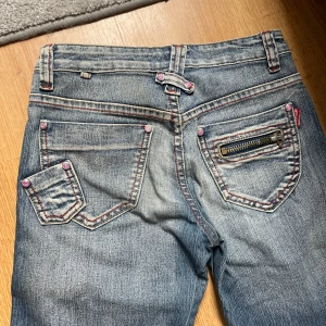Blå bootcut jeans från Kat Jeans - Snygga blå bootcut jeans från Kat Jeans med slitna detaljer, rosa sömmar och nitar med rosa stenar. Jeansen har dragkedja, dekorativ dragkedja på bakfickan och breda ben. Använd, jättefina bakfickor, har lite smuts innuti men annars perfekt. Kan gärna sänka priset, bara postnord!!! Kan skicka den 02 aug och framåt