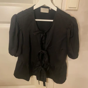Svart blus med knyt från ADPT - Svart blus från ADPT med tre dekorativa knyt framtill och puffiga korta ärmar. Blusen har en lätt v-ringning och volang nedtill, vilket ger en trendig och söt look. Perfekt för dig som gillar detaljer och en feminin stil.