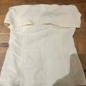 Offwhite axelbandslös tubtopp i linne - Jätte fin tubbtopp som ger en clean stil och fräsch look till vad som. Grejen man ska kunna haka fast på för att underlätta att dra upp dragkedjan har gått sönder men inget fel på dragkedjan. Säljer för att den är för liten💕