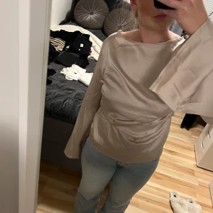 Beige satintopp från Boohoo - Snygg beige satintopp från Boohoo med lös passform och långa vida ärmar. Toppen har en slät, glansig yta och rynkade detaljer i midjan som ger en cool siluett. Båtringad halsringning och elegant fall. Perfekt för dig som gillar stilrena och trendiga plagg. Strl 40 men upplevs som 36/38. Endast testad.