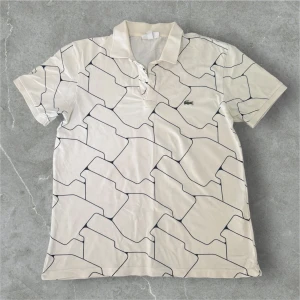 Vit pikétröja från Lacoste med mönster - Snygg vit pikétröja från Lacoste med svart grafiskt mönster över hela tröjan.                                                Färg: Vit & Svarta Linjer (Se bilderna för bästa färgätergivning) Storlek: Medium (M) Inte 100% säker pà exakt storlek men uppskattas passa som en SMALL (vänligen dubbelkolla mätten för säkerhets skull) Mătt: • Axel till axel: 46 cm • Längd: 64 cm Skick: Mycket fint skick - Visar inte märke av användning. Inga häl eller större skador. Material: 100% Bomull Ord pris: 1350kr