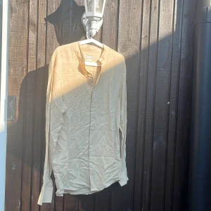 Beige linneskjorta från Zara - Snygg beige skjorta från Zara i linne med regular fit. Skjortan har knappar framtill, lång ärm och en stilren, kraglös henley-modell. Perfekt för dig som gillar en avslappnad men ändå trendig look.