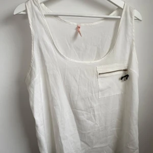 Vitt linne från Sweewë Paris - Snygg vit linnetopp från Sweewë Paris med rund halsringning och ärmlös design. Toppen har en bröstficka med dragkedja och en liten svart dekoration som detalj. Perfekt för en fräsch och clean look under varma dagar.