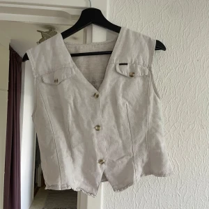 Ljusbeige väst - Snygg väst i jeans- liknande material med v-ringning och knappar. Färgen är så fin, blandning av vit, grå, beige. Västen har två bröstfickor med och figursydda sömmar. Perfekt att styla över en t-shirt eller bära som den är. Köpt second hand. 
