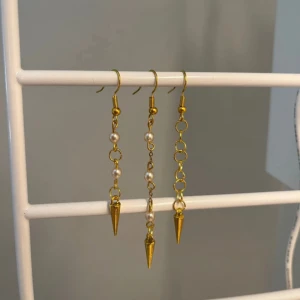 Guldiga örhängen med nittar och pärlor - Säljer coola guldiga örhängen med lång kedja, små vita pärlor och spetsig kon längst ner. Unik design som sticker ut och passar dig som gillar edgy detaljer. Säljs i par men mixa gärna för en asymmetrisk look. 1 för 19kr, 2 för 35kr och alla för 45kr! Handgjort! Kontakta mig om du vill köpa - eller två. Om du vill köpa alla tryck på köp nu!