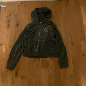 Grön velourjacka med huva från Lindex - Mysig grön velourjacka från Lindex med huva och dragkedja. Jackan har långa ärmar, ribbade muddar och ett glittrigt S-märke framtill i strass. Perfekt för dig som gillar en chill och snygg stil.