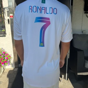 Al Nassr Ronaldo CR7 vit fotbollströja - Säljer en vit Al Nassr fotbollströja med CR7-tryck och Ronaldo 7 på ryggen. Tröjan är köpt utomlands men kvaliten och materialet är 10/10. Helt oanvänd ! 