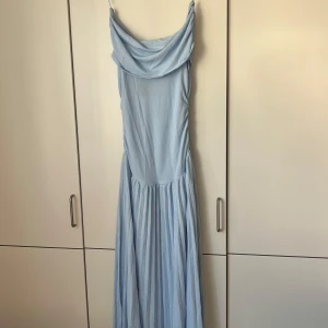 Ljusblå långklänning med draperad urringning - Superfin ljusblå långklänning med smala axelband och draperad urringning. Klänningen har en flowy kjol och är perfekt för sommarens alla tillfällen. Materialet känns lätt och svalt, och modellen är riktigt elegant och trendig.
