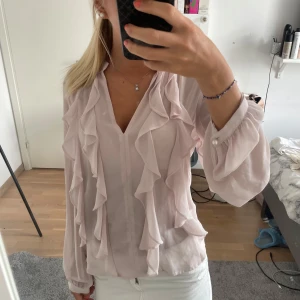 Ljusrosa blus med volanger - Supersöt ljusrosa blus med v-ringning och långa, luftiga ärmar. Blusen har flera volangdetaljer framtill som ger en romantisk vibe. Tunt och flowy material, perfekt för dig som gillar en feminin och trendig stil.
