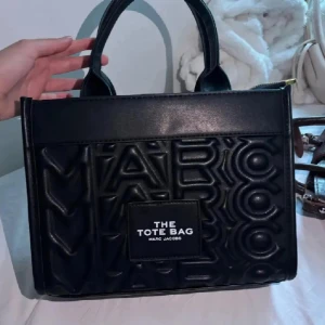 Svart tote bag från Marc Jacobs - Svart handväska från Marc Jacobs med präglad logotyp över hela väskan och tydlig patch framtill med texten 'The Tote Bag'. Väskan har två stadiga handtag och är tillverkad i syntetskinn. 💕