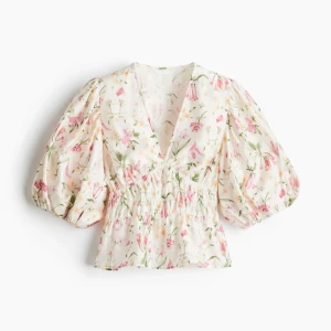 Blommig blus med puffärm och v-ringning - Supersöt blommig blus i vitt med rosa och gröna detaljer. Den har v-ringning, smockad midja och puffiga trekvartsärmar. Blusen är croppad och har volang nertill, perfekt för en fräsch och somrig look.