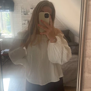 Vit offshoulder blus från Gina Tricot - Superfin vit blus från Gina Tricot med offshoulder-modell och långa, puffiga ärmar. Blusen har en rynkad struktur och volangdetalj upptill. Perfekt att styla med jeans eller kjol för en trendig look.