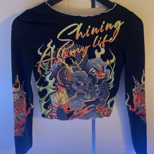 Svart långärmad topp med draktryck - Cool svart långärmad topp med färgstarkt drakmotiv, eldflammor och texten 'Shining All my life' i neonfärger. Toppen är croppad och har tajt passform, perfekt för dig som vill sticka ut med edgy streetstyle.