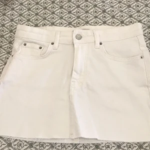 Vit jeanskjol från Gina Tricot - Snygg vit jeanskjol från Gina Tricot med klassisk femficksdesign, bälteshällor och rå kant nertill. Kjolen har knapp och dragkedja framtill samt en läderpatch med logga bak. Perfekt för en fräsch och stilren look.