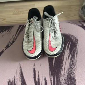 Nike Phantom GT2 fotbollsskor vit/rosa - Nike Phantom GT2 fotbollsskor i vitt med svarta och rosa detaljer. Skorna har snörning, rund tå och en ovandel i syntetmaterial med textur för bättre bollkontroll. Snygg design med rosa Swoosh och PHANTOM GT2-tryck på sidan. Perfekta för dig som vill sticka ut på planen.