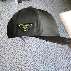 Svart keps från Prada  - Snygg svart keps från Prada med klassisk böjd skärm och ikonisk metallogo framtill. Kepsen är tillverkad i glansig nylon och har justerbar rem baktill för perfekt passform. En stilren accessoar som lyfter din outfit.