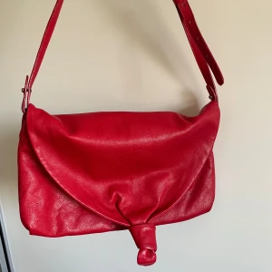 Röd axelväska i skinn från Think! - Snygg röd axelväska i mjukt skinn från Think! med justerbart axelband och unik knutdetalj framtill. Väskan har dragkedjeficka på baksidan och flera fack inuti för smart organisering. Perfekt för dig som vill ha färg till outfiten💗💗