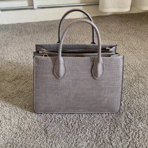 Grå handväska med croco-mönster - Snygg grå handväska i croco-mönstrat skinnimitation med guldiga detaljer och dragkedja. Väskan har både handtag och en avtagbar, bred axelrem i svart och vitt. Perfekt storlek för vardagens essentials och ger en clean, trendig look.