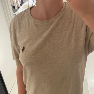 Beige t-shirt från Polo Ralph Lauren - Beige t-shirt från Polo Ralph Lauren med klassisk rund hals och korta ärmar. T-shirten har den ikoniska broderade hästloggan på bröstet och är gjord i mjuk bomull. Enkel och stilren modell som passar till allt.