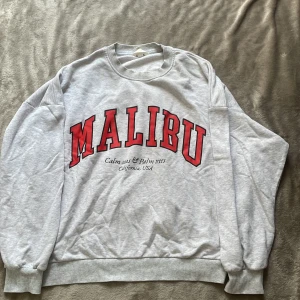 Grå sweatshirt med Malibu-tryck H&M - Grå sweatshirt från H&M med stort rött 'MALIBU'-tryck på bröstet och svart text under. Tröjan har rund hals, ribbade muddar och är långärmad. Perfekt för en chill och avslappnad stil.