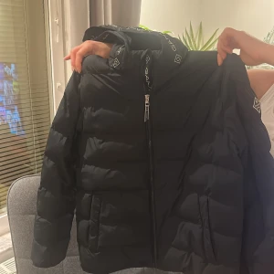 Svart pufferjacka från GANT (priset kan diskuteras vid snabb affär) - Snygg svart pufferjacka från GANT med logga på ärmen och insidan av kragen. Jackan har dragkedja framtill, hög krage och två fickor. Perfekt för kalla dagar och riktigt stilren med diskreta detaljer. Helt i nyskick.