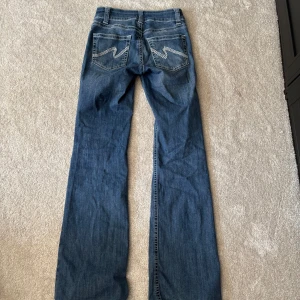 Blå bootcut jeans från Nelly - Snygga blå jeans från Nelly med bootcut passform och klassisk femficksdesign. Jeansen har låg midja, dubbla knappar och coola detaljer på bakfickorna. Tillverkade i jeansmaterial med lätt slitning nertill för en avslappnad vibe. 