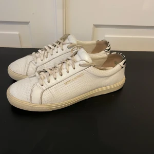 Vita Saint Laurent sneakers i skinn - Snygga vita sneakers från Saint Laurent i perforerat skinn med diskret guldlogga på sidan. Skorna har snörning och en cool detalj med svartvitt zebramönster på hälen. Perfekta för en clean och trendig look.