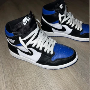 Nike Air Jordan 1 High svart/blå/vit - Säljer ett par Nike Air Jordan 1 High i svart, blått och vitt skinn. Klassisk siluett med vit sula, svarta swoosh och vita snören. Perfekta för dig som vill sticka ut med en ikonisk sneaker med streetkänsla.