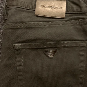 Svarta byxor från Emporio Armani - Snygga svarta byxor från Emporio Armani med klassisk femficksdesign och ikonisk logga i metall på bakfickan. Byxorna har en rak passform och är tillverkade i ett mjukt bomullsmaterial med lite stretch för extra komfort. Perfekta för en clean och stilren look. Köptes för 1999kr