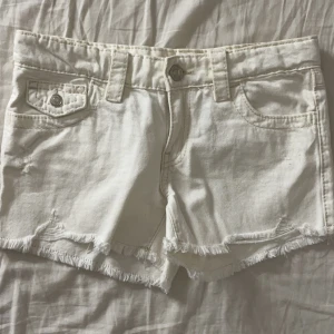 Vita jeansshorts från Gina Tricot - Snygga vita jeansshorts från Gina Tricot med fransig kant och coola fickdetaljer med knappar både fram och bak. Klassisk femficksmodell och bälteshällor. Perfekta för varma dagar och en avslappnad stil.