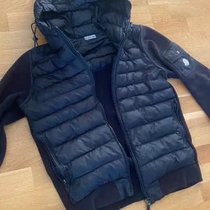 Svart Moncler pufferjacka med huva - Svart pufferjacka från Moncler med quiltad framsida och huva. Jackan har ribbade muddar, dragkedja framtill och en ficka med Moncler-logga på ärmen.