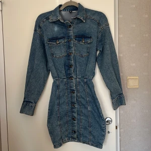 Jeansklänning från H&M Divided - Snygg jeansklänning från H&M Divided i klassisk blå denim. Kort modell med långa ärmar, markerad midja och knappar hela vägen framtill. Två bröstfickor med lock och krage ger en cool, avslappnad look. Perfekt för dig som gillar denimvibbar! 