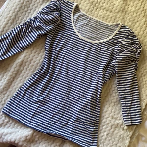 Randig trekvart topp, blå/vit - Snygg randig topp i blått och vitt med trekvartsärm. Rund halsringning med vit kant. Perfekt för dig som gillar en klassisk men trendig stil. Mjuk och skön bomullskänsla, passar året runt.