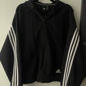 Svart Adidas zip kofta - Svart hoodiejacka från Adidas med ränder längs ärmarna och ryggen. Oversized passform. Perfekt för dig som gillar sportig och avslappnad stil.