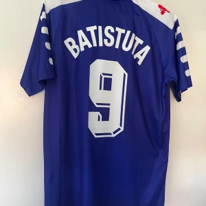 Fiorentina Batistuta 90-tals fotbollströja - Klassisk blå Fiorentina fotbollströja från Fila med Nintendo som sponsor och Batistuta 9 på ryggen. Tröjan har korta ärmar, vit krage med tunn rand, klubbmärke på bröstet och vita detaljer på axlar och ärmar. Perfekt för dig som älskar retro och italiensk fotboll.