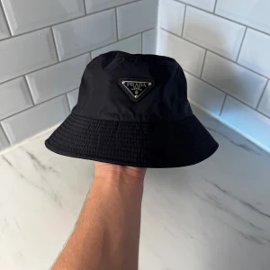 Svart bucket hat från Prada - Stilren svart bucket hat från Prada med ikonisk triangellogo framtill. Hatten är tillverkad i glansig syntet och har klassisk form med bred, nedåtlutande brätte. Perfekt accessoar för en trendig streetstyle-look.