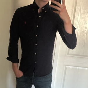 Mörkblå skjorta från Ralph Lauren - Säljer en mörkblå slim fit skjorta från Ralph Lauren med klassisk krage och vita knappar. Skjortan har lång ärm och den ikoniska röda Polo-loggan broderad på bröstet. Perfekt för dig som gillar stilrena och tidlösa plagg.