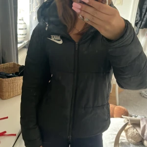 Svart pufferjacka från Nike, S - Säljer en svart pufferjacka från Nike i storlek S. Jackan har huva, dragkedja framtill och en clean design med Nike-logga på bröstet. Perfekt för kalla dagar när du vill hålla stilen och ändå vara varm.