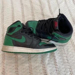 Nike Air Jordan 1 GS pine green svart EUR 36 - Nike Air Jordan 1 GS hightop i colorway PINE GREEN (svart version). Skorna har svart snörning, gröna detaljer på tå, sula och häl samt röd Nike Air-logga på plösen. Perfekt för dig som gillar streetstyle och ikoniska sneakers.