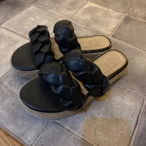 Svarta flätade sandaler - Snygga svarta sandaler med två breda, flätade remmar i skinnimitation och platåsula i repstil. Perfekta för en trendig look under varma dagar. Sulan är platt och ger en avslappnad vibe.