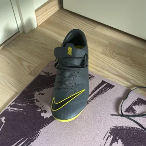 Nike Mercurial grå/gula fotbollsskor - Säljer ett par Nike Mercurial fotbollsskor i grått med gula detaljer och logga. Skorna har snörning och är tillverkade i syntetmaterial för bästa grepp och känsla på planen. Snygg design med tydliga gula linjer och Mercurial-tryck på hälen.