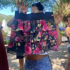 Blommig volangkjol med resårmidja - Supersöt kort kjol med flera volanger och blommigt mönster i rosa, gult och svart. Kjolen har bred resår i midjan och är perfekt för dig som gillar färg och rörelse. Lätt och luftig känsla, passar till sneakers eller sandaler.