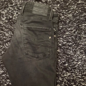 Svarta Replay Anbass jeans, stl 31 - Säljer ett par svarta Replay Anbass jeans i storlek 31. Jeansen har klassisk femficksdesign, snygga detaljer på bakfickan och en svart läderpatch i midjan. Slimmad passform och tillverkade i mjukt jeansmaterial med stretch för extra komfort.