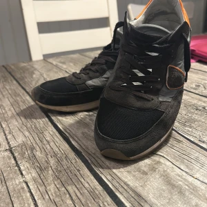 Svarta sneakers med orange detalj - Snygga svarta sneakers från Philippe Model med grå och orange detaljer. Ovandel i mocka och textil, med klassisk snörning och vit sula. Skorna har en cool kontrast med orange häl och diskret logga på sidan. Perfekta för dig som gillar streetstyle och unika detaljer.